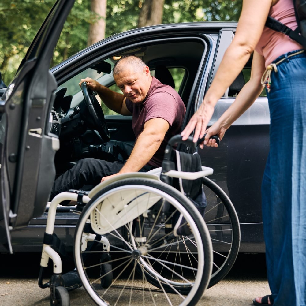 A caregiver assists a person with a disability as they transition from a car to a wheelchair.